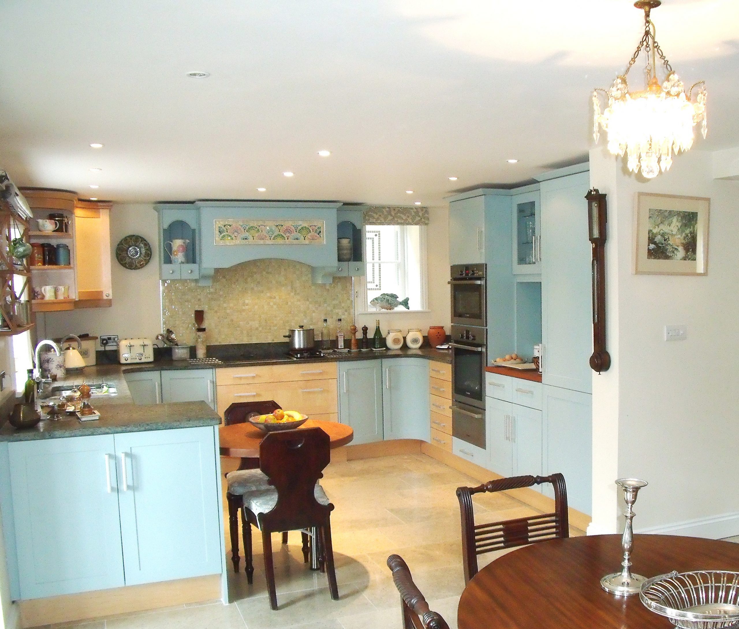 Bright kitchen with pastel blue cupboards, wooden worktops, and beige tiled splashback. This kitchen's design features a small island with fruit, built-in ovens, sink under a window, dining table and chairs, wall clock, and chandelier overhead. | Saxon Kitchens | Andover, Hampshire