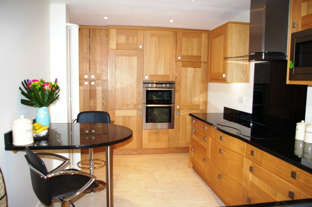 Modern kitchen design with light wood cupboards, black worktops, built-in oven and microwave, black glass hob, bar stools at a small black dining table, and a vase of colourful flowers. Worktop has jars and fruit; the space is bright and tidy. | Saxon Kitchens | Andover, Hampshire